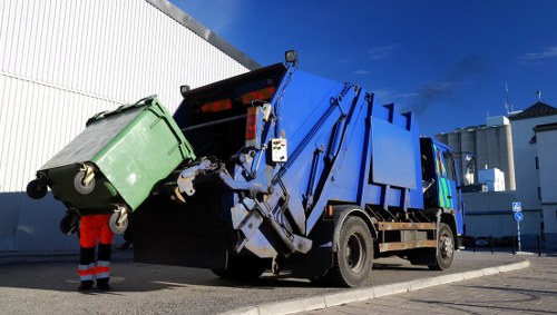 A large blue industrial rubbish collection truck with a hydraulic lifting mechanism at the rear is in the process of emptying a green wheeled waste bin onto the truck's sorting area. The bin, made of plastic with a textured surface, features a black lid and is positioned near the left side of the image. It is partially raised, tilted, and suspended above the open hopper of the truck, demonstrating the waste collection process. The scene is set outdoors on a paved area adjacent to a white industrial building with vertical paneling, under a clear blue sky. A worker dressed in red high-visibility clothing and safety gloves stands close by, overseeing the operation. The truck has black tires, visible reflective markings, and is parked on a street or driveway that appears to be in an urban or commercial district, potentially within or near Wanstead or surrounding areas. The environment is well-lit with natural daylight, emphasizing clean, professional waste management activities conducted by Office Clearance Wanstead, which offers rubbish removal services in the local area.