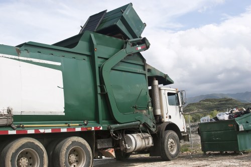 A large commercial waste collection truck with a white cab and dark green compactor body is parked outdoors on a gravel surface near a green waste container filled with mixed rubbish, including plastic bags and other refuse. The truck's compactor mechanism is raised, and the hydraulic system appears ready for unloading or collection. The background features a partly cloudy sky with patches of blue, rolling green hills, and distant rocky outcrops, suggesting a rural or semi-rural location. The vehicle’s tires are rugged and designed for heavy-duty use, and the scene is illuminated by natural daylight, emphasizing the metallic textures and colors of the truck and waste container. This image is relevant to rubbish removal services offered by Office Clearance Wanstead, serving areas within or near E11 postcode, combining professional waste collection with the scenic landscape typical of the Wanstead region.