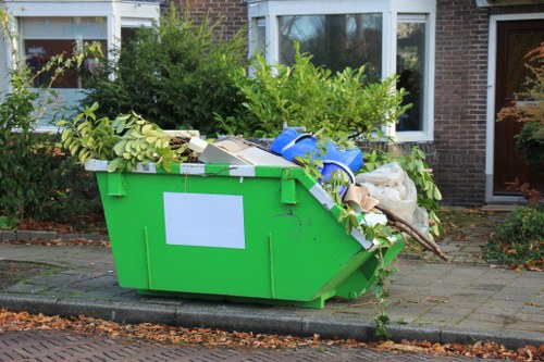 A large, bright green industrial skip positioned on a paved driveway in front of a residential building with brick walls and double-glazed windows. The skip is filled with a variety of waste materials, including green leafy branches and twigs, white cardboard boxes, plastic blue wrapping, and some white and beige fabric or paper items. The waste appears to be a mix of garden debris and general rubbish. The skip is situated immediately next to a curb, with a small patch of pavement and the residential garden visible in the background, featuring bushes and trees. The environment suggests a collection or removal of garden waste and household rubbish, likely managed by a local rubbish removal service such as Office Clearance Wanstead, based in the Wanstead area, on the outskirts of London. The scene is captured in natural daylight, highlighting the textured surfaces of the waste and the clean, bright finish of the skip.