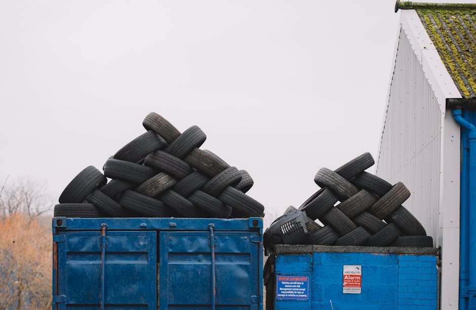 A large blue skip filled with numerous used black rubber tires stacked in an overlapping manner, with some tires leaning at various angles. The tires appear weathered, with worn treads and slight disc