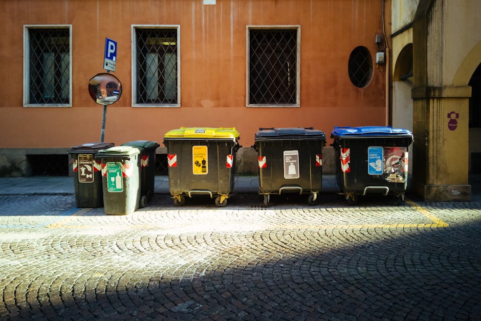 A row of five large wheeled rubbish bins lined up on a cobbled street in front of an orange-toned building with three barred windows. The bins vary in color—green, yellow, black, black with a white