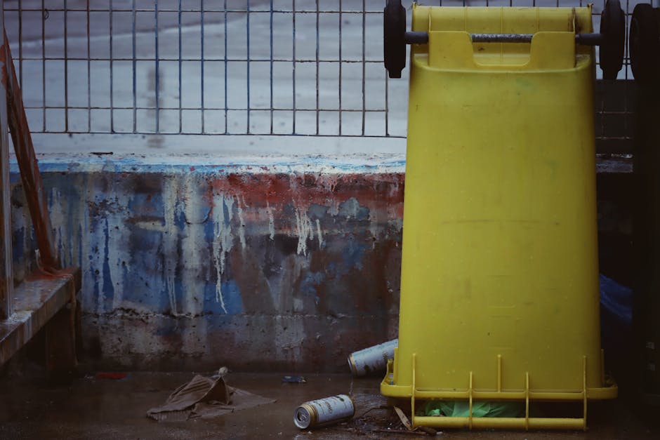 A row of five large wheeled rubbish bins lined up on a cobbled street in front of an orange-toned building with three barred windows. The bins vary in color—green, yellow, black, black with a white
